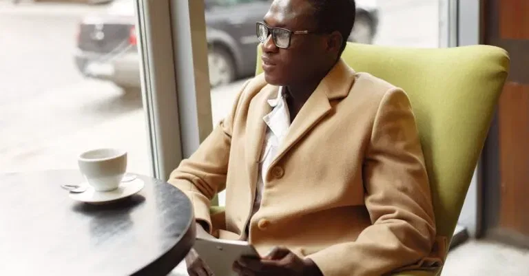 Serious African American Male In Trendy Formal Suit And Eyeglasses Sitting On Cozy Chair In Cafe With Cup Of Coffee And Browsing Tablet