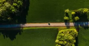 Aerial Shot Of A Car Driving On A Tranquil Road Through Lush Green Countryside On A Sunny Day.