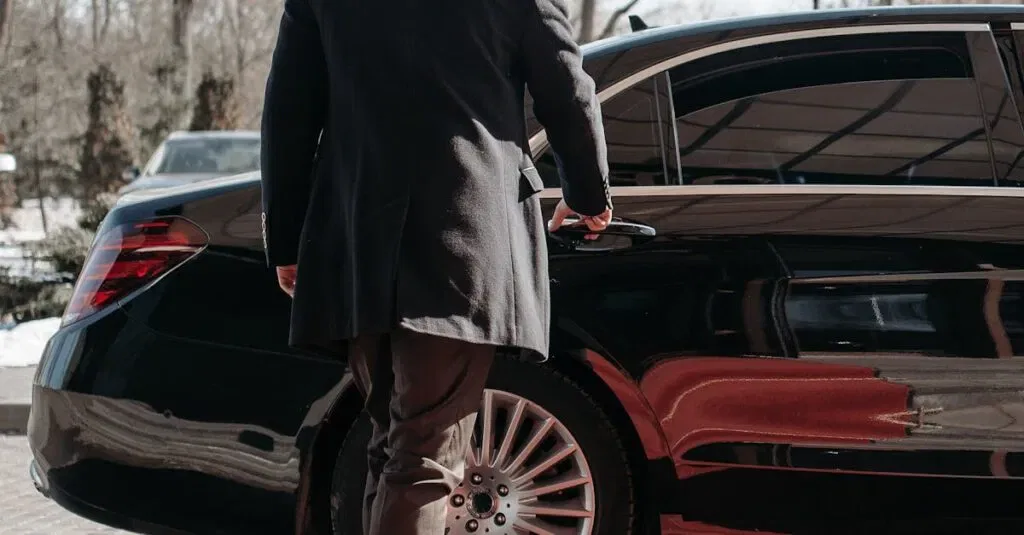 Bald Man In A Black Suit Approaches A Luxury Black Car On A Sunny Day.