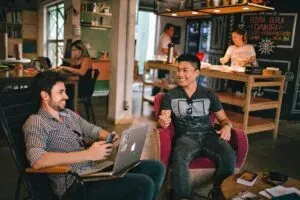 Two Men Enjoying A Relaxed Conversation With Laptops In A Cozy Brazilian Café.