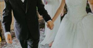A Romantic Moment Capturing A Bride And Groom Holding Hands In Elegant Wedding Attire Indoors.
