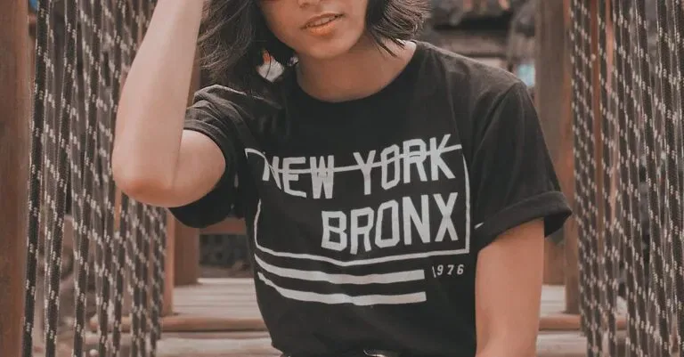 Confident Woman In Glasses Wearing New York Bronx Shirt Sitting On A Rustic Wooden Bridge Outdoors.