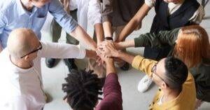 A Diverse Group Of Professionals Joins Hands In A Team-Building Exercise Indoors.