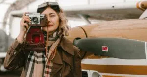 Fashionable Woman With Vintage Camera Posing By An Airplane In An Open Hangar.