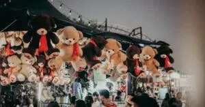 A Bustling Carnival Scene At Night Featuring Teddy Bear Prizes And A Lively Crowd.