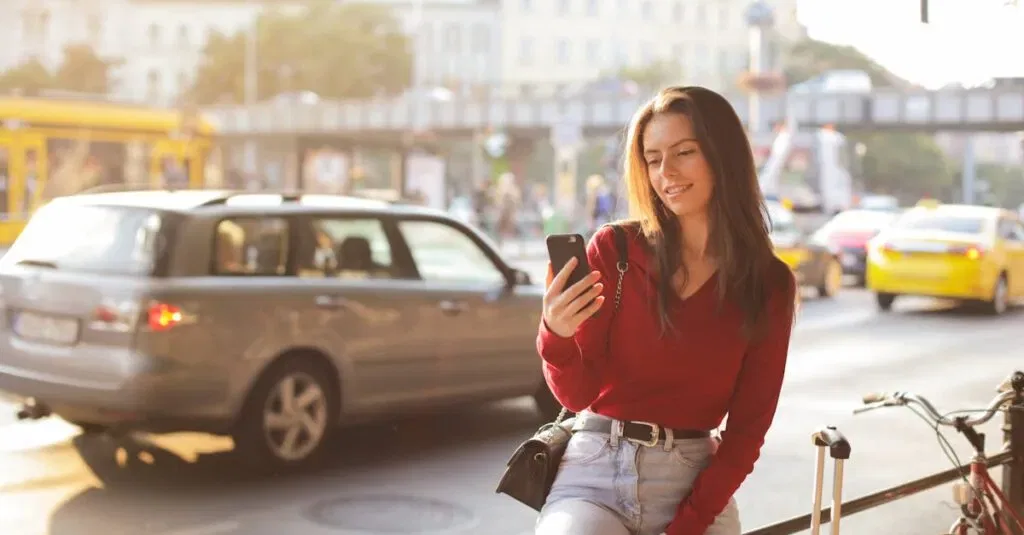 Smiling Woman In A Red Sweater Uses Her Phone On A Sunny Urban Street. Cars And Bicycles Surround Her.