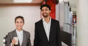 Two Smiling Professionals Walking In A New York Subway Station, Holding Coffee And Briefcase.