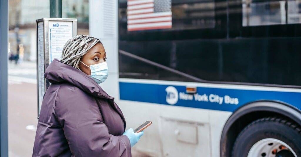 Woman With Mask And Mobile Device Waiting At New York City Bus Stop During Pandemic.