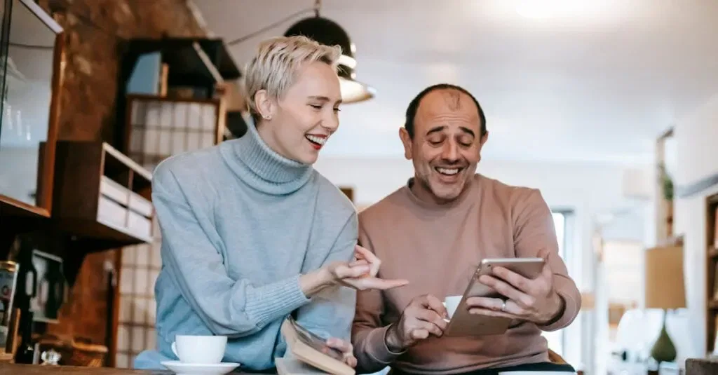 Cheerful Adult Diverse Couple In Casual Clothes Watching Funny Video On Tablet And Laughing While Drinking Coffee At Table In Apartment