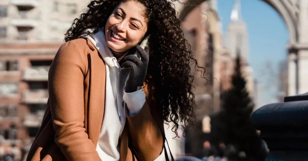 Young Woman With Curly Hair Happily Chatting On Phone In A City Setting, Enjoying A Sunny Day.