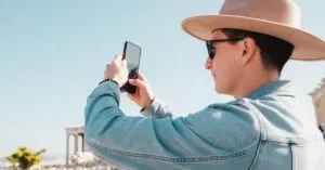 Man In Denim Jacket Taking Photo In Front Of Ancient Columns, Capturing His Travel Memories.