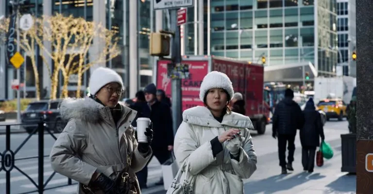 Two Women In Winter Coats Walking On A Bustling City Street With Holiday Lights.