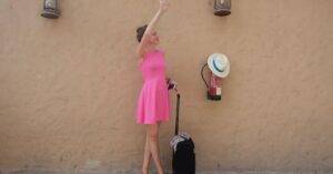 A Woman In A Pink Dress With Luggage, Waving For A Cab On A Sunny Day In Dubai.