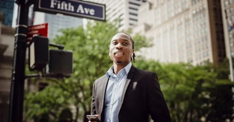 From Below Of Young Handsome Black Male In Smart Suit Listening To Music And Waiting For Car To Pass While Standing Near Street Sign In Manhattan On Sunny Day