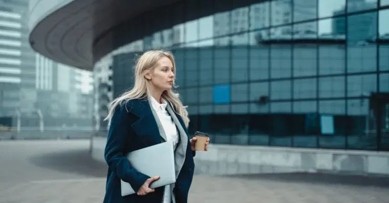Confident Businesswoman Walking Outside Modern Office Building Holding Laptop And Coffee.