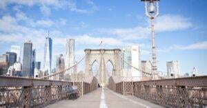 Wide-Angle View Of The Brooklyn Bridge Leading To New York City'S Iconic Skyline Under Blue Skies.