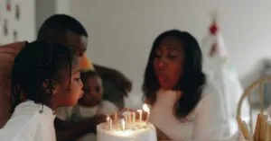 A Joyful Family Celebrating A Birthday By Blowing Out Candles On A Cake Indoors.