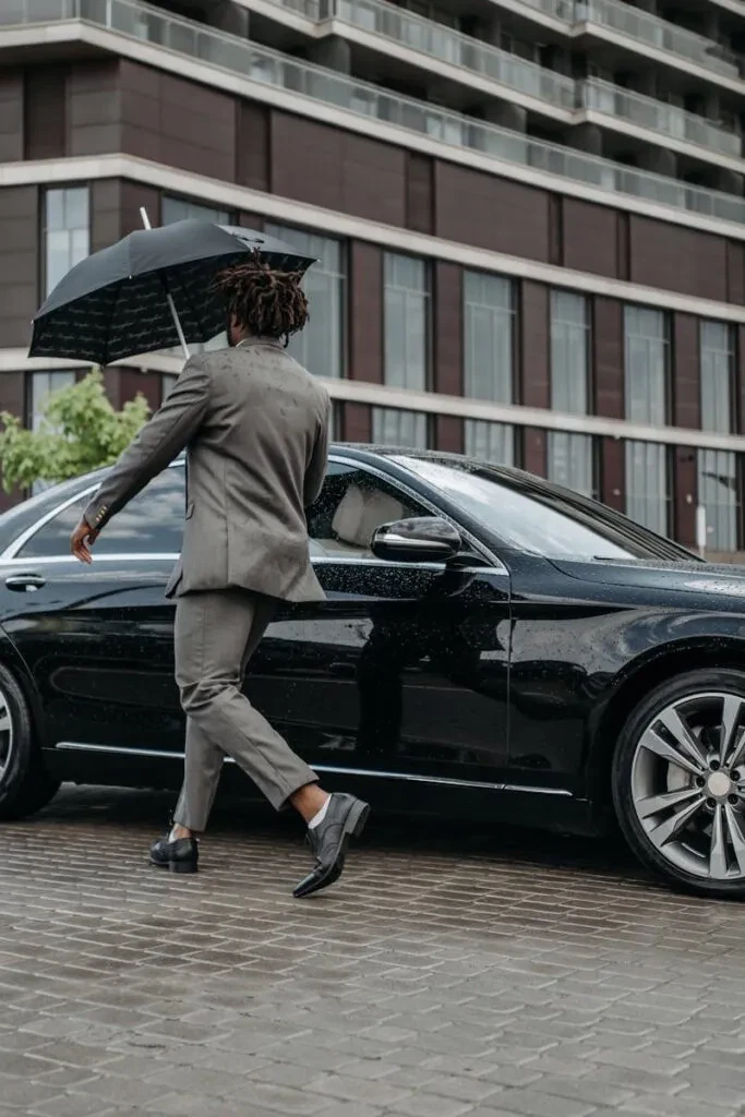 Man Holding Umbrella Opens Car Door On Rainy Day In Front Of Modern Building.