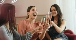 Three Women Laughing And Clinking Champagne Glasses In A Joyful Indoor Celebration.