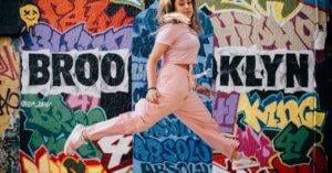 Corporate Travel Articles &Amp; News 6 May 20, 2026 Energetic Woman In Pink Jumpsuit Jumping Against A Vibrant Brooklyn Mural, Capturing Urban Street Art Culture.