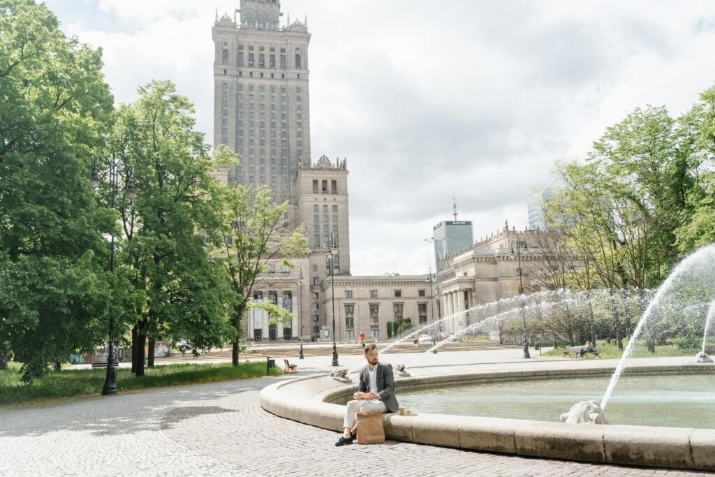 A Man Sits By A Fountain Near The Palace Of Culture And Science In Warsaw, Enjoying A Casual Lunch Outdoors.