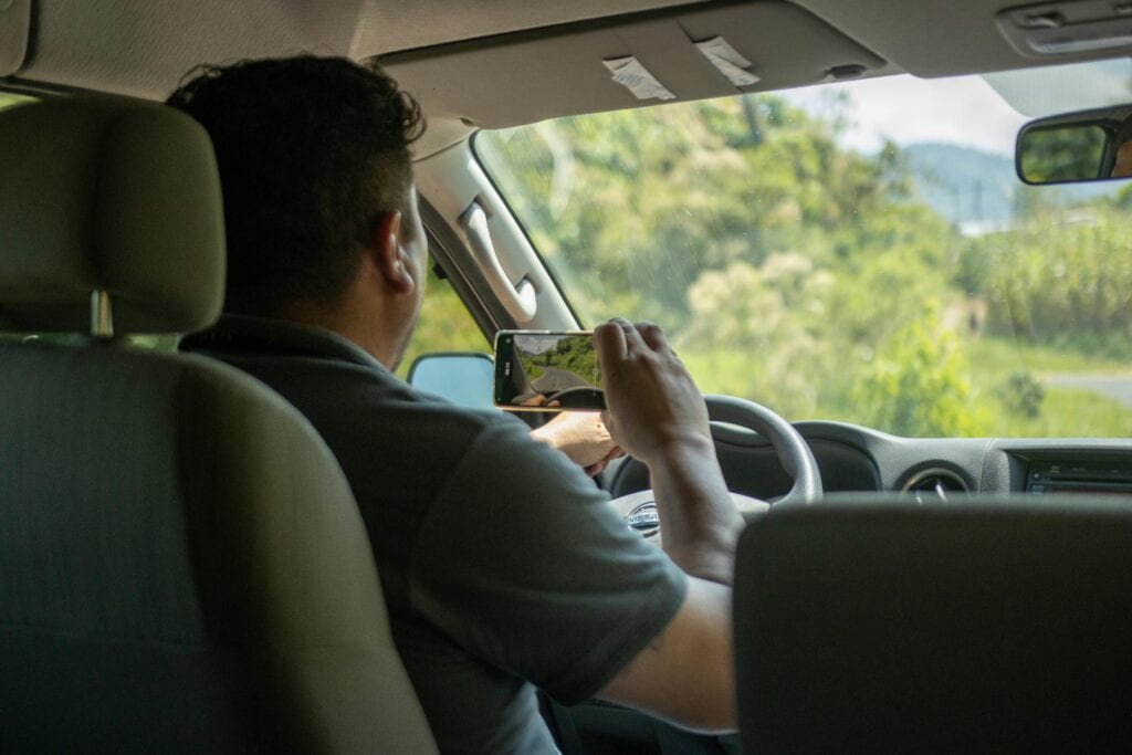 A Man Uses A Smartphone To Capture A Scene Outside While Driving In A Car Interior During The Day.