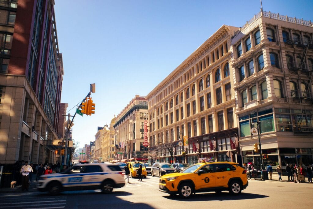 Dynamic Street View Of A Bustling New York City Intersection With Taxis And Police Cars Under Sunny Skies.