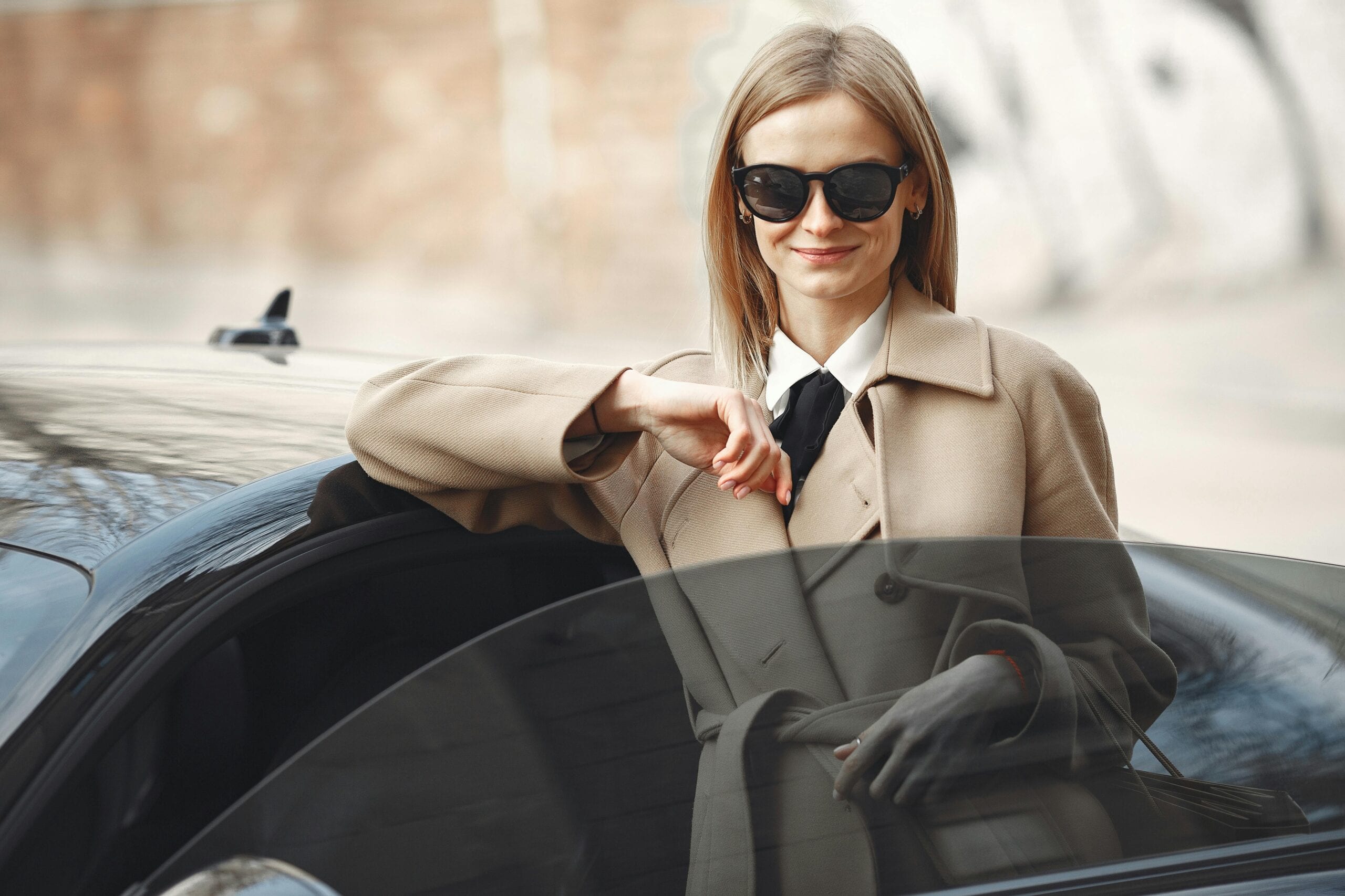 Smiling Female Driver In Stylish Coat And Sunglasses Leaning On Automobile While Standing On City Street With Paper Bags
