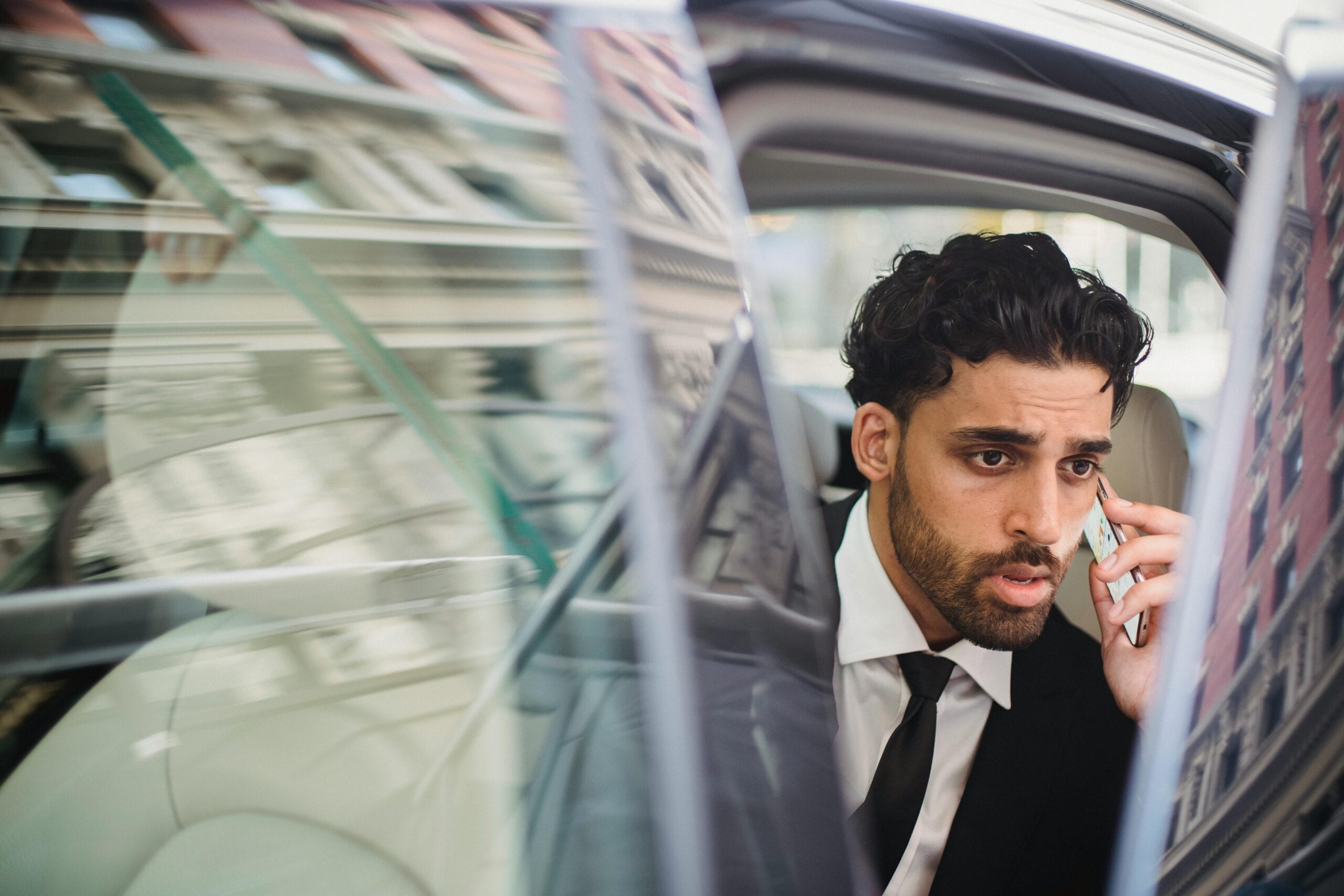 A Professional Man In A Suit Talking On His Phone While Seated In A Car With An Open Door.