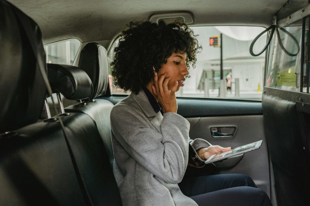 Side View Of African American Woman In Stylish Outfit Sitting On Passenger Seats Of Taxi And Talking With Wireless Earphones While Browsing Tablet