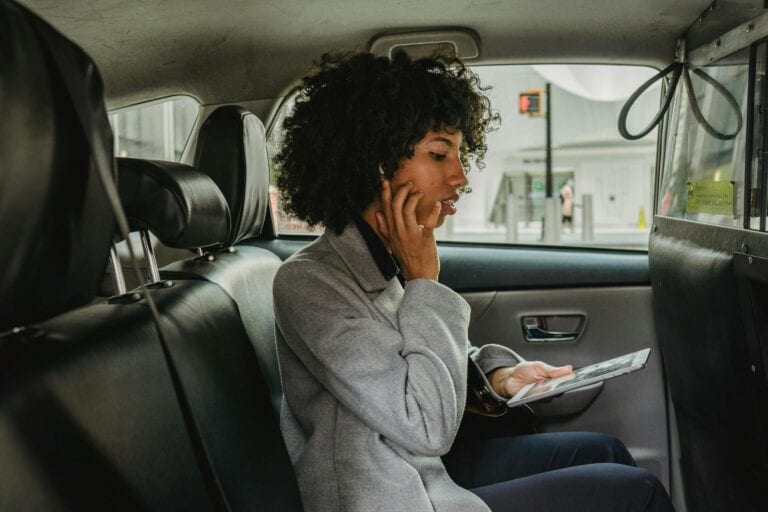 Side View Of African American Woman In Stylish Outfit Sitting On Passenger Seats Of Taxi And Talking With Wireless Earphones While Browsing Tablet