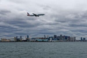 Passenger Plane Flying Over Water Towards Boston Skyline, Cloudy Day.