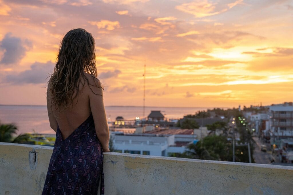 A Woman With Long Hair Gazes At A Vibrant Sunset From A Rooftop, Creating A Serene And Picturesque Moment.