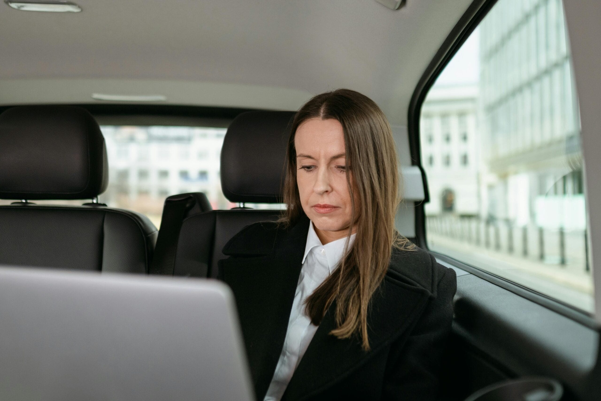 Focused Businesswoman In Formal Attire Working On A Laptop In The Back Seat Of A Car.