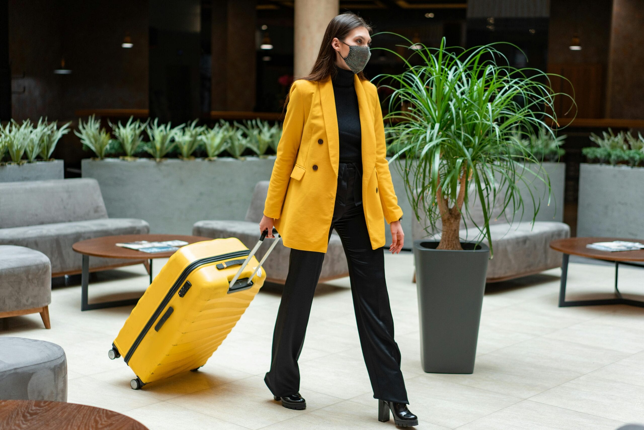 Fashionable Woman In Yellow Blazer And Face Mask Walking With Suitcase Indoors.