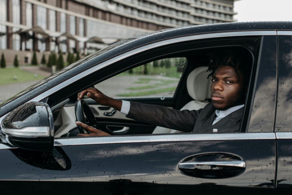 African American Man Confidently Driving A Sleek Black Luxury Car Outside A Modern Building.