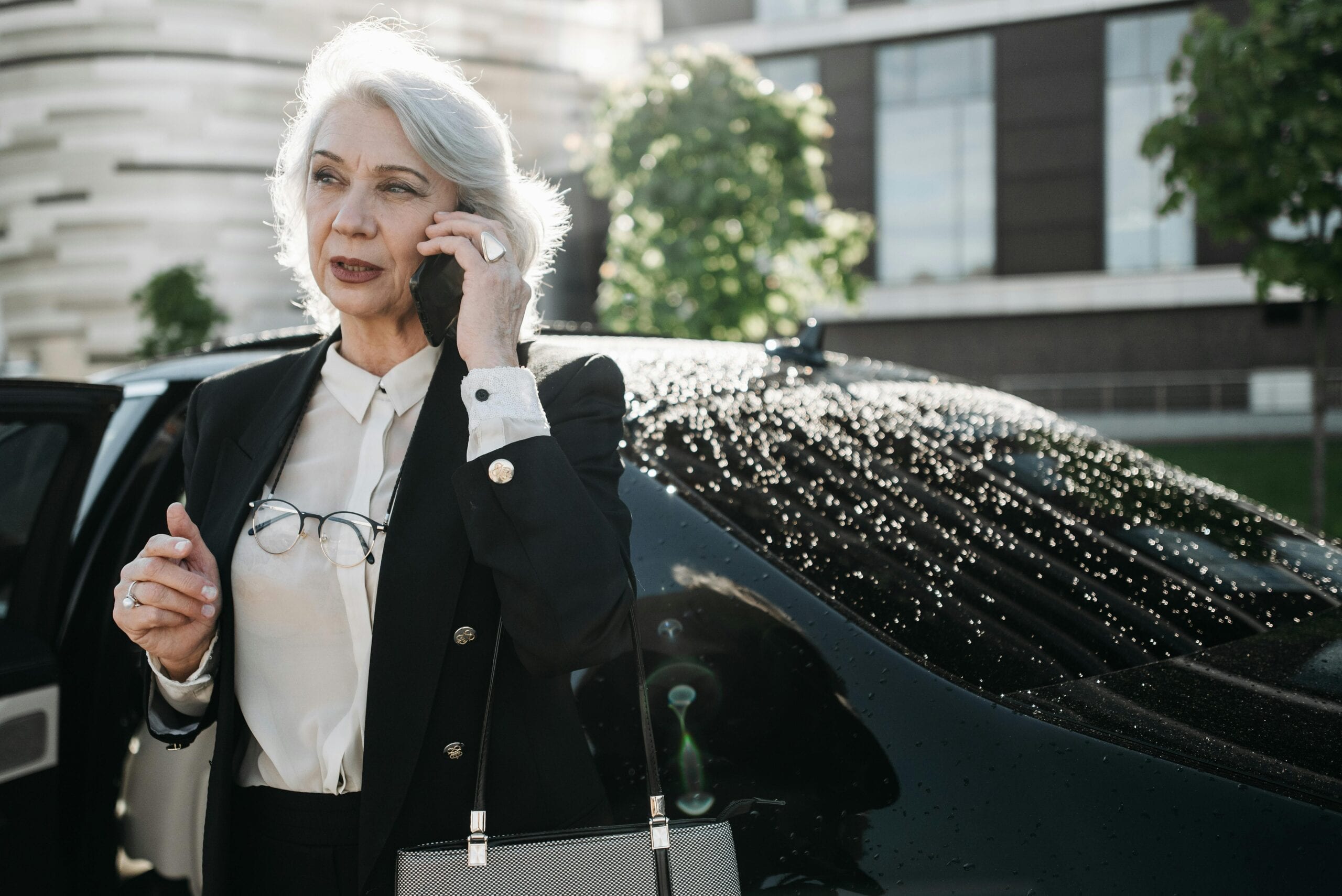 Senior Woman In Formal Attire Using A Mobile Phone Near A Black Car Outdoors.