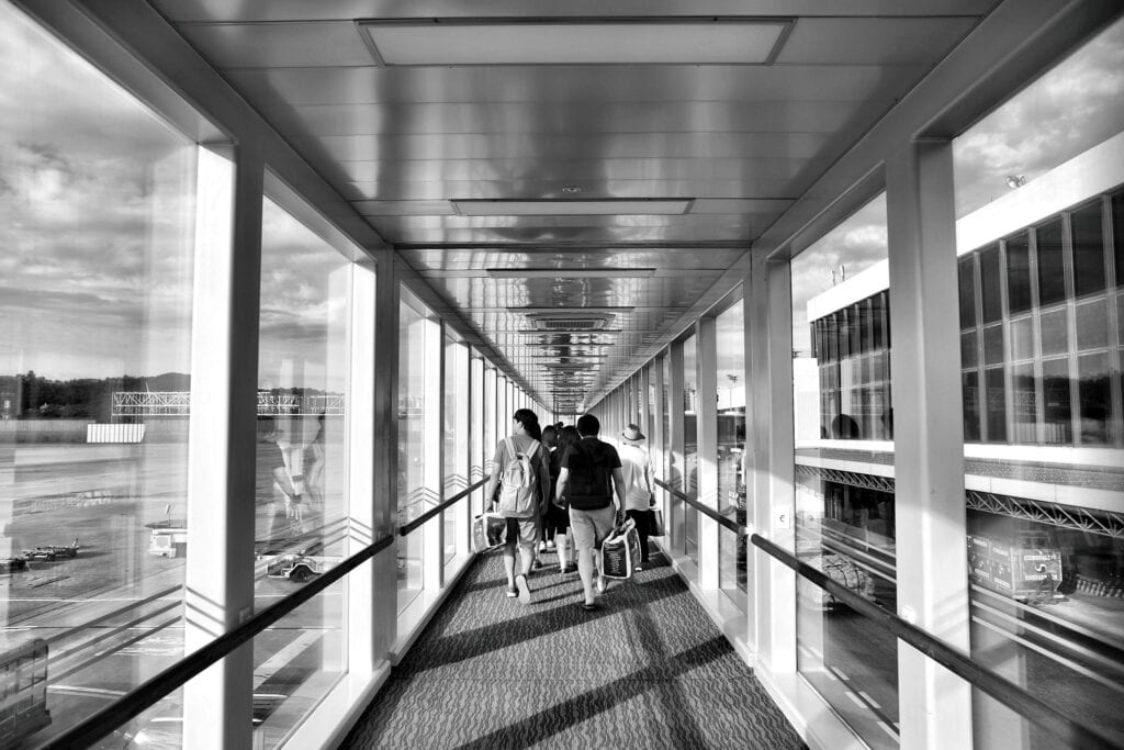 Travelers Walking Through A Glass Corridor At An Airport Terminal In Black And White.