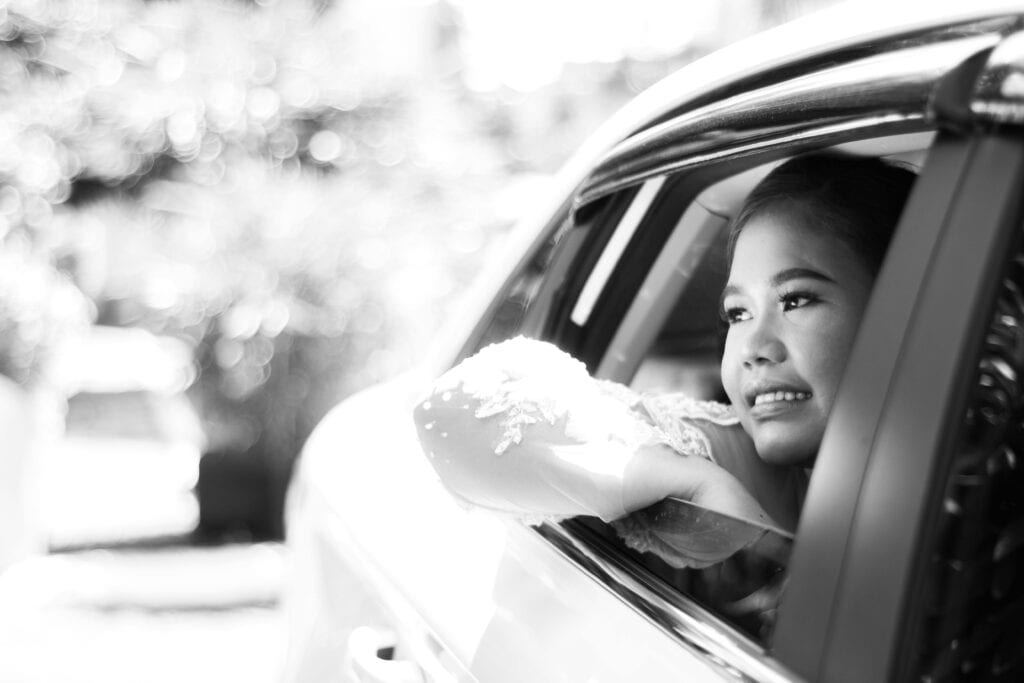 Black And White Photo Of A Smiling Bride In A Wedding Dress, Looking Out The Car Window With Joy.