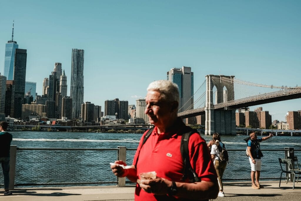Tourists Enjoy The View Of The Brooklyn Bridge Against The New York City Skyline On A Sunny Day.
