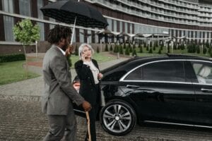 New York City (Nyc) Article &Amp; News 6 May 19, 2026 Two Business Professionals With A Luxury Car Outdoors, Using An Umbrella And Smartphone.