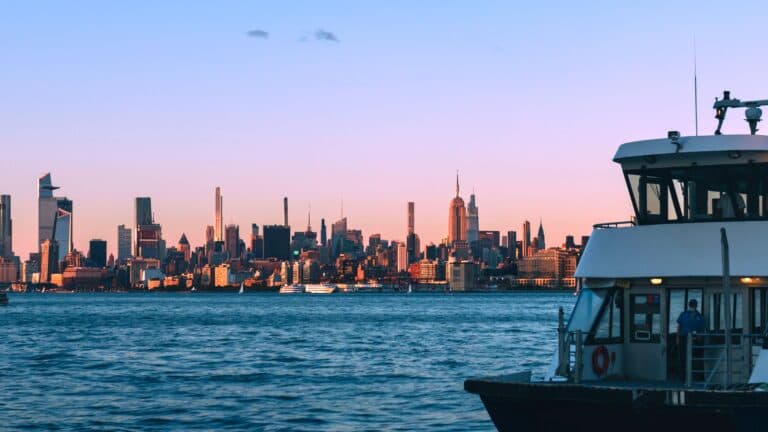 View Of New York City'S Skyline During Dusk With A Ferry In The Foreground, Capturing Urban And Water Elements.