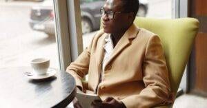 Serious African American Male In Trendy Formal Suit And Eyeglasses Sitting On Cozy Chair In Cafe With Cup Of Coffee And Browsing Tablet