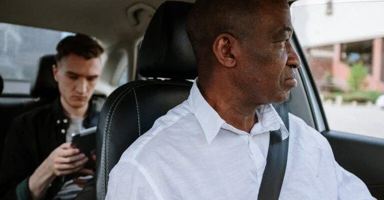 A Professional Driver In A White Shirt Navigates With A Young Passenger Using His Phone In The Backseat.