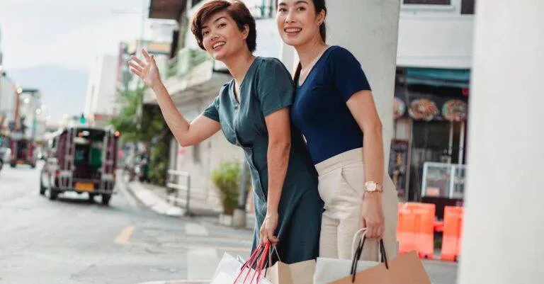 Low Angle Of Smiling Female Friends With Paper Bags Catching Cab After Shopping In Mall