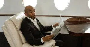An Adult Man In A Black Suit Reads A Newspaper While Sitting In A Private Jet'S Luxurious Interior.