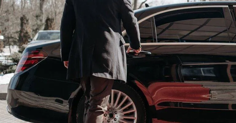Bald Man In A Black Suit Approaches A Luxury Black Car On A Sunny Day.