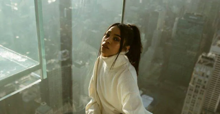 Young Woman In Cozy Sweater Posing By A Glass Wall Overlooking New York City From A Skyscraper.