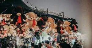 A Bustling Carnival Scene At Night Featuring Teddy Bear Prizes And A Lively Crowd.