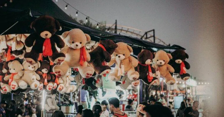 A Bustling Carnival Scene At Night Featuring Teddy Bear Prizes And A Lively Crowd.
